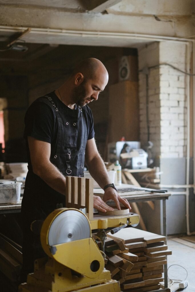 Side view of skilled carpenter polishing round piece of wood while working with belt and disc sander in workshop with stack of wood