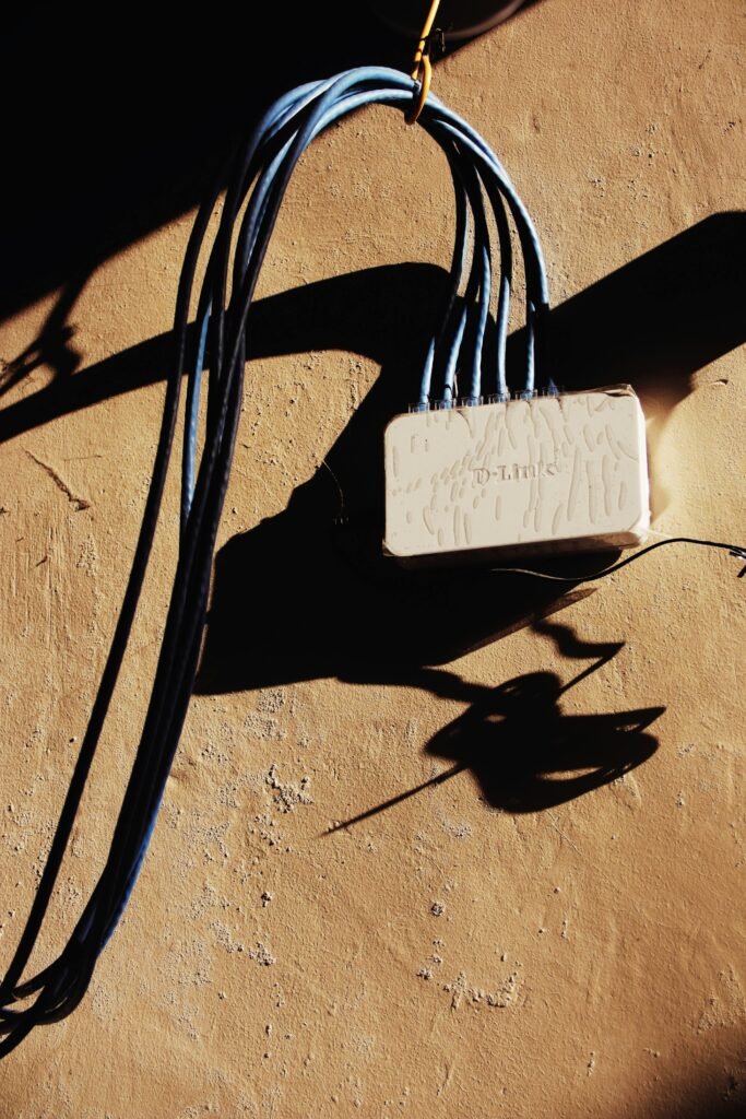 White electrical network adapter with various blue wires hanging on rough brown wall of building with bright sunlight and shadow